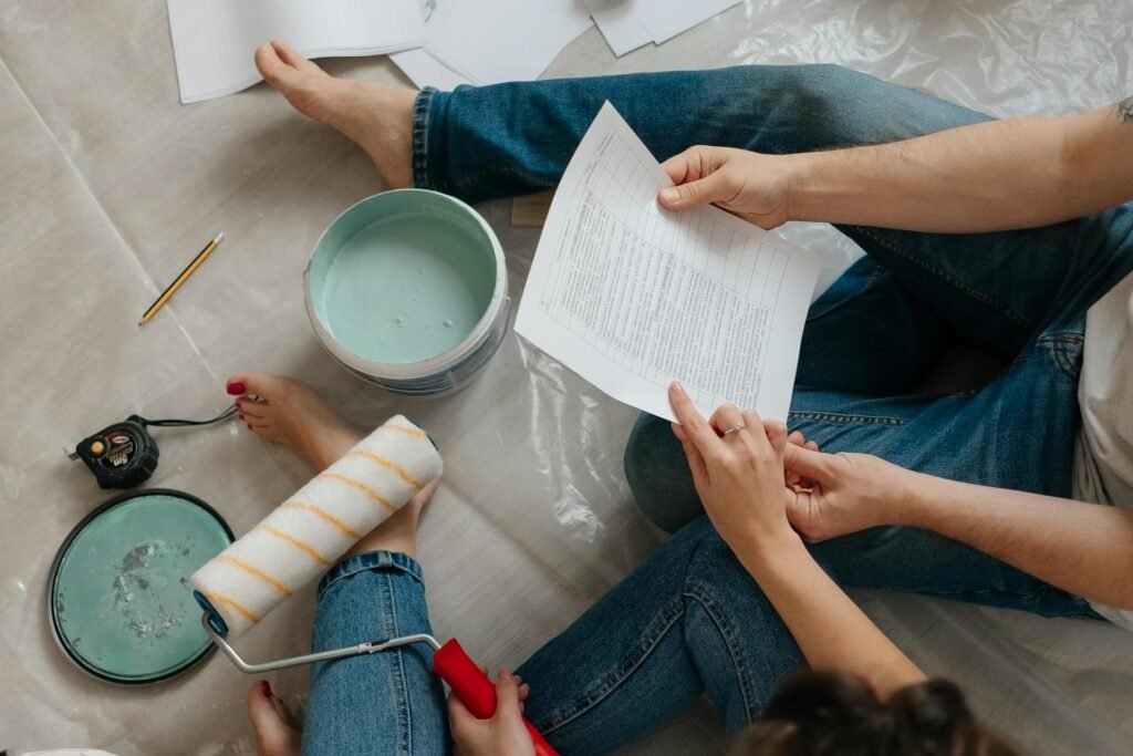 Two people in jeans sit on a drop cloth reviewing a document while surrounded by light blue paint, a roller, and a tape measure.