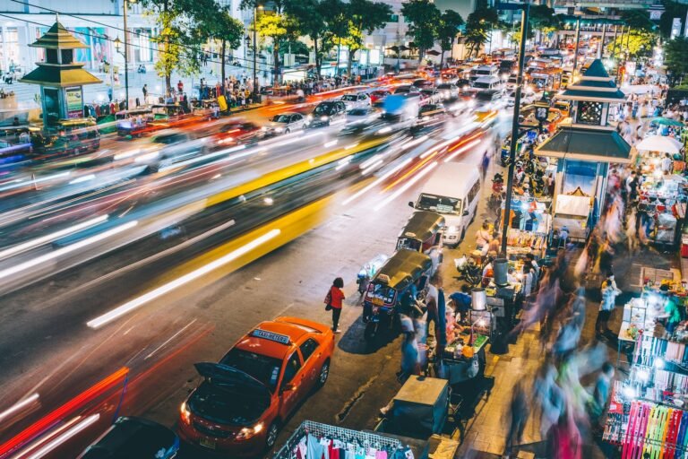 High-angle night view of a bustling city street with light trails from fast-moving traffic contrasting with blurred pedestrians and street vendors along the roadside.
