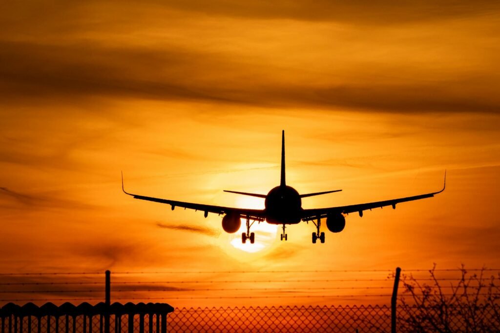 Silhouette of a commercial airplane landing against a brilliant orange sunset sky, seen over a chain-link fence.