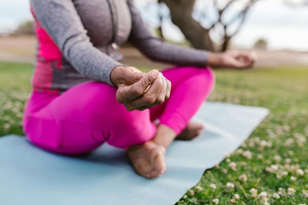 Close-up of an older person in bright pink leggings meditating in a cross-legged position outdoors on a yoga mat with their hands in a mudra.