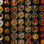 An overhead view of numerous small bowls containing various Asian soups, desserts, and savory items arranged in neat rows alongside plates of waffles and segmented wooden boxes of fruit.