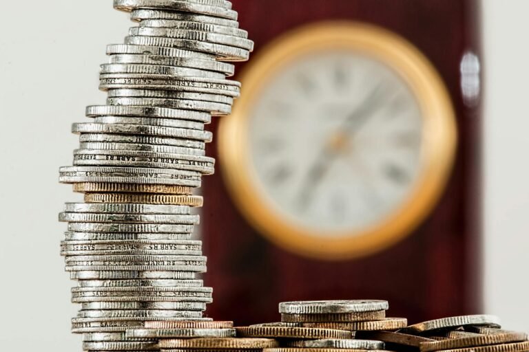 Tall stack of silver and gold coins sharply focused in the foreground with a blurred gold-rimmed clock in the background, symbolizing time and money.
