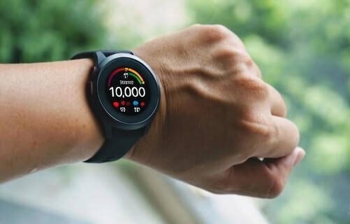 Close-up of a wrist wearing a black smartwatch displaying a fitness goal of 10,000 steps against a blurred green outdoor background.